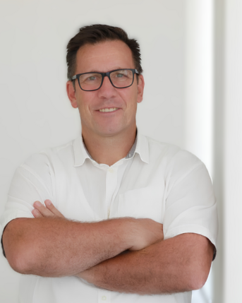 Mann mit Brille und kurzem Haar, der in einem weißen Hemd steht und die Arme vor der Brust verschränkt. Helle, neutrale Umgebung.